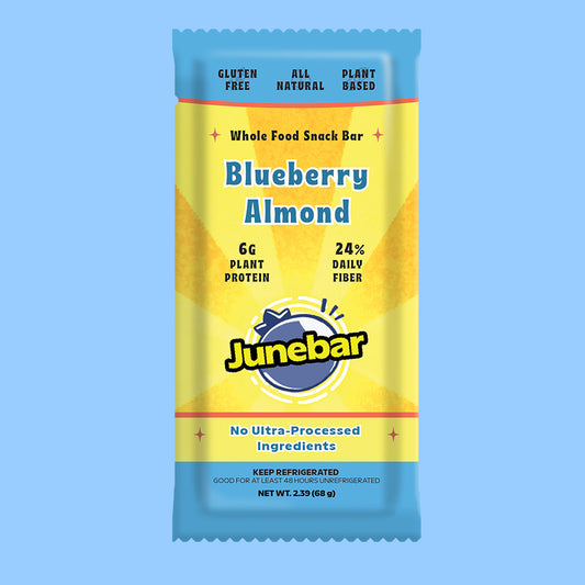 A Junebar Blueberry Almond whole food snack bar in its wrapper, shown against a solid light blue background. The wrapper is primarily yellow with blue text and a blue brand logo, with small red accent lines.