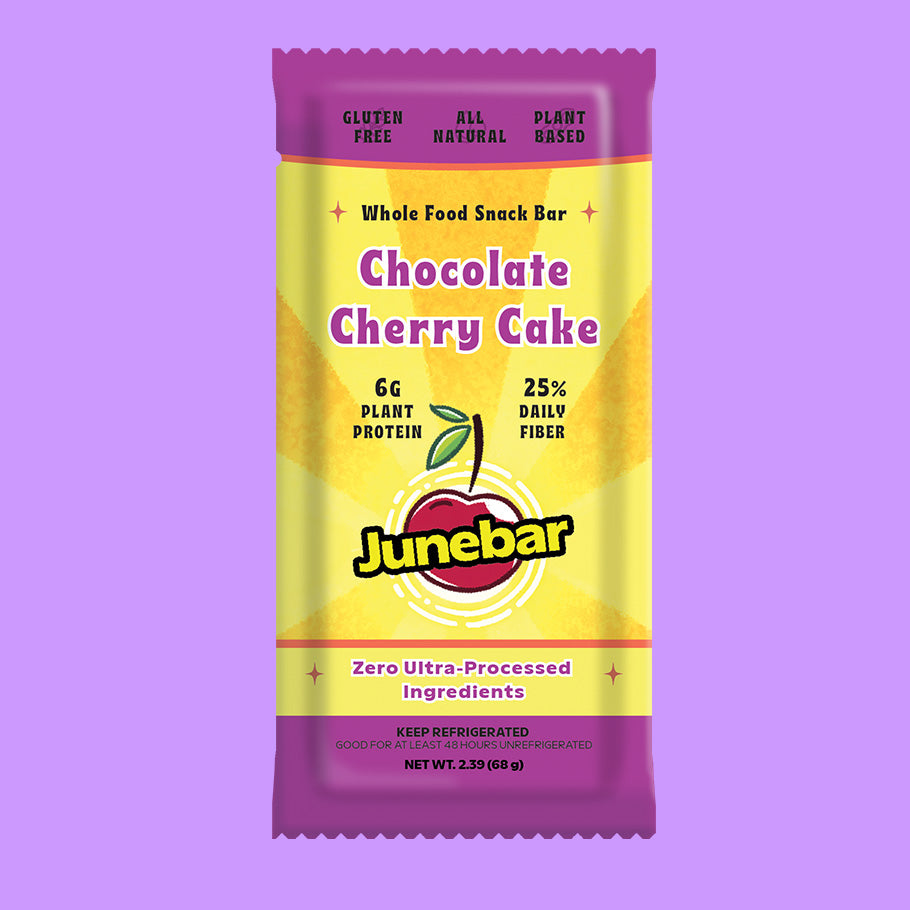 A packaged Junebar Chocolate Cherry Cake snack bar centered on a solid lavender background. The wrapper is primarily yellow with purple top and bottom bands, and the Junebar logo is prominently displayed near the bottom.