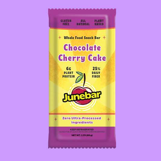 A packaged Junebar Chocolate Cherry Cake snack bar centered on a solid lavender background. The wrapper is primarily yellow with purple top and bottom bands, and the Junebar logo is prominently displayed near the bottom.